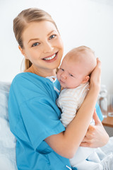happy young mother sitting on bed with adorable baby and smiling at camera in hospital room
