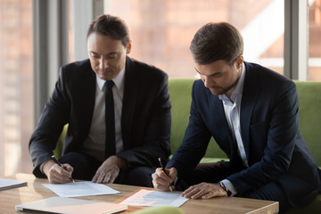 Two successful middle aged businessmen in suits signing contracts for services at meeting,...