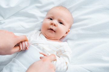 cropped shot of mother holding hands of adorable infant baby lying on white bedding