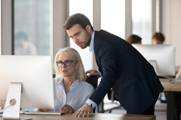 Serious male mentor manager ceo supervising middle aged female employee helping with computer problem explaining online work teaching older colleague learning use new software in corporate office.