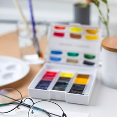 Watercolor paints, brushes and drawing paper on a white table by the window next to glasses, flower vases and a lamp. Hobby time