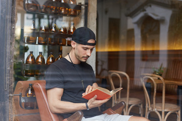 Thoughtful unshaven man reads books, learns some tips for successful project, sits at modern cafe, dressed in casual clothing, focused on reading, has dark stubble. Leisure and literature concept.