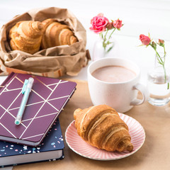 Croissant and a cup of cocoa on a white desktop next to note pads. Break concept