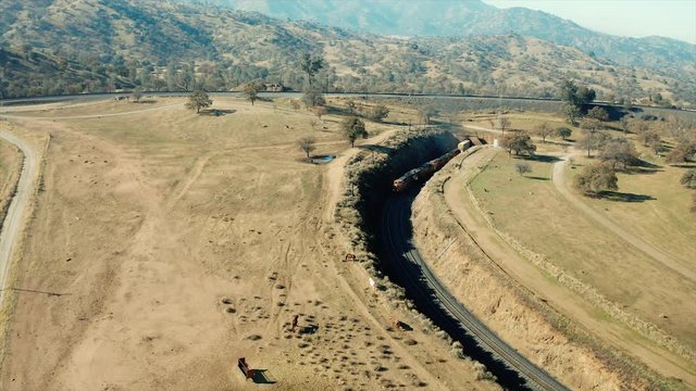 The aerial camera flying in reverse 250 feet overhead of FOUR powerful diesel locomotives pulling a very long freight train coming out of the northern tunnel of the Tehachapi Railroad Loop.