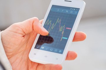 Close up finger hand of a trader touching smartphone screen with trading interface dashboard with candlestick chart in mobile trading forex or stock application