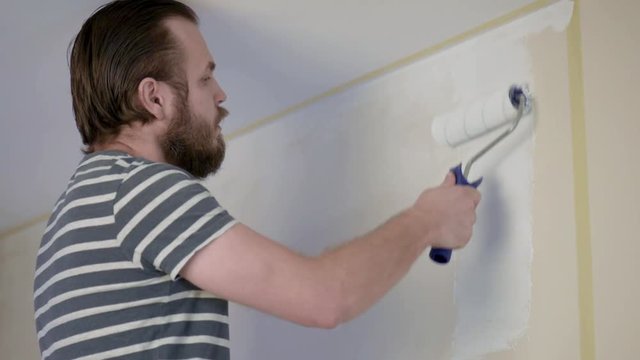 Bearded Man Painting interior Walls at Flat using Paint Roller. Handsome young man doing Repairs - actively paints wall with white paint. Home renovation or Redecoration concept.
