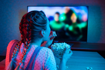 A young girl watching movies and eating popcorn with a bowl on the background of the TV. The color bright lighting, blue and red. Relax, rest at home when watching TV, film. Background for design - 246840961