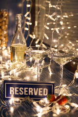 Closeup strawberry, two crystal glasses, bottle champagne sparkling wine, reserved nameplate on wooden table, window, garlands. Concept date evening in restaraunt, 8 march, Valentine's Day, christmas