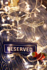 Closeup strawberry, two crystal glasses, bottle champagne sparkling wine, reserved nameplate on wooden table, window, garlands. Concept date evening in restaraunt, 8 march, Valentine's Day, christmas