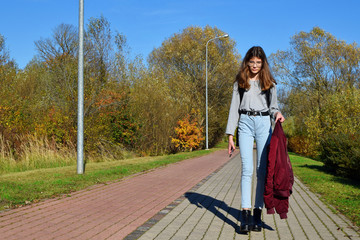 Teen girl wearing glasses and jeans walking pavement pathway from school to home in beautiful autumn day landscape.