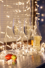 Closeup fresh strawberries, two crystal glasses, bottle of champagne sparkling wine on background of window, garlands, bokeh. Concept cozy evening in michelin restaurant, dating, dinner, Valentine Day