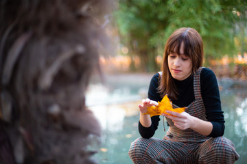 Pensive attractive lady holding yellow maple leaf near water in garden on blurred background