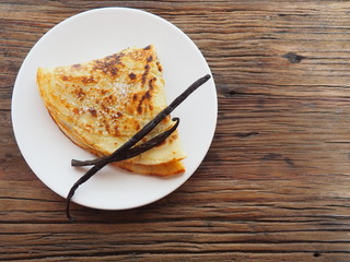 Traditional french crepe (or pancake) on a white plate, with sugar and vanilla pods on top, dark brown wood background