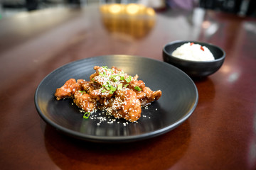 Sweet and Sour Chicken with sesame and green onions on the black plate, small bowl of rice behind it. Red wood table