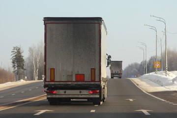 Logistics, international freight by road -  dirty white euro semi trucks drive next to the two-lane asphalted highway road in the winter day, back view