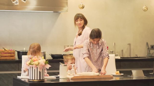 A family of four cooks all together. They prepare food for lunch. Kids are busy at the table and mother checks on them from time to time.