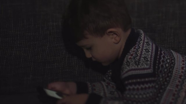 Adorable boy sitting on the sofa in the living room and playing with gadget. Child learning how to use smartphone. Boy texting on the phone. Technology and lifestyle concept . Blurred movie 