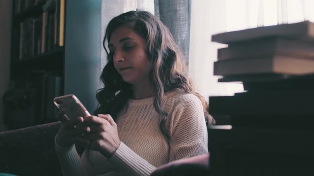 Brunette millenial woman is on her phone in her library, gets a text and smiles