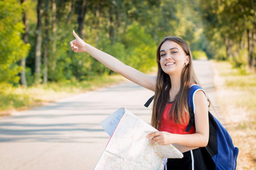 Naklejka premium Smiling girl traveling hitchhiking putting up a hand tries to stop the car. Traveler girl with heavy backpack and a map in her hands wants to stop a car to get a lift to a destination