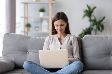 Happy teen girl using laptop sitting on couch at home, young woman surfing web on notebook, female doing online shopping, making order on website, chatting with friend or working studying in internet