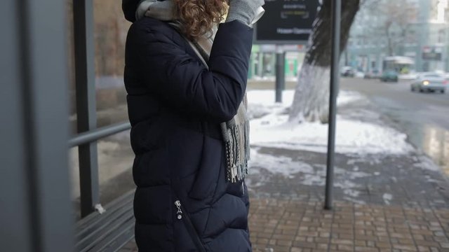 Unrecognizable young woman dressed in warm clothes waiting for bus at stop on winter day, drinking coffee and looking at wristwathces.
