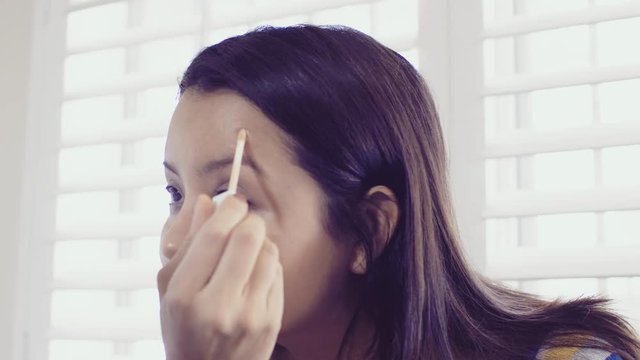 Hispanic millenial woman applies cover up / concealer to a blemish in her bright white bathroom