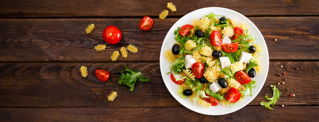 Italian pasta salad with fresh tomato, cheese, lettuce and olives on wooden background. Mediterranean cuisine. Cooking lunch.  Healthy diet food