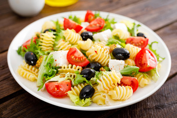 Italian pasta salad with fresh tomato, cheese, lettuce and olives on wooden background. Mediterranean cuisine. Cooking lunch.  Healthy diet food