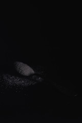 A dark photo of a spoon with white sugar on a black stone background with copy space, horizontal view, low key lighting