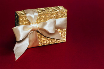 Beautiful shiny golden present box with a silk bow on deep red background. Copy space.