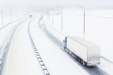Trucks with cargo trailers on the highway in a snow storm. Top view of a snowy asphalt road in bad weather conditions with poor visibility and slippery surface. Transportation in a winter season