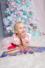 Fototapeta premium Portrait of cute european little blonde princess girl with crown in beautiful dress lying on the floor and reading book in decorated studio in christmas location with toys and christmas tree