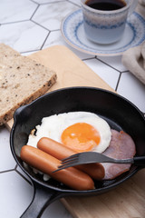 Breakfast fried egg with sausage and ham on a iron pan served with whole grain bread in the morning