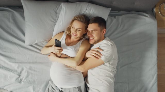 Loving Young Couple Spending Morning in Bed, Pregnant Young Woman Shows Her Partner Something on a Touchscreen Smartphone. Searching for Baby Clothes, Browsing Through Social Media, Posting Pictures.