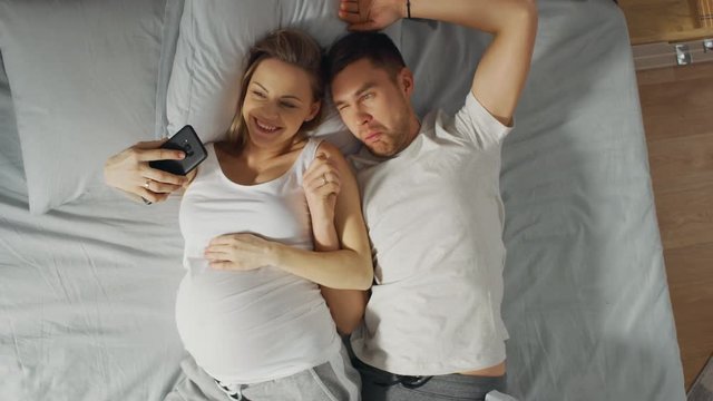 Loving Young Couple Spending Morning in Bed, Pregnant Young Woman Shows Her Partner Something on a Touchscreen Smartphone, Taking Selfie and Sharing Picture on Social Networks.