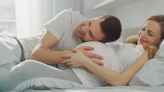 Happy Young Couple Cuddling Together in the Bed, Young Woman is Pregnant, Loving Partner Leans and Listens to a Baby Moving and it's Heartbeat in the Belly. Tender and Loving Moments of Family Life.