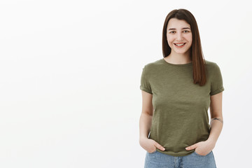 Waist-up shot of good-looking bright and sincere brunette female with short hairstyle and friendly smile grinning at camera holding hands in pockets in casual relaxed pose over gray wall