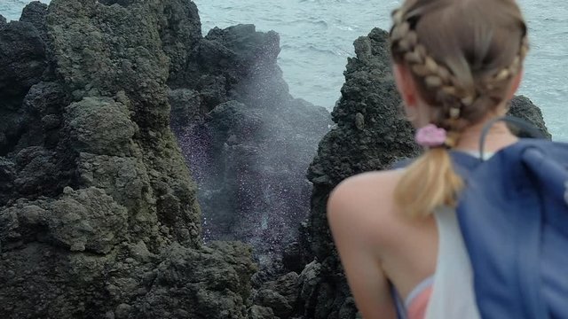 Caucasian lady hiker watches on the rocks with blow hole and its powerful spray, Maui, Hawaii