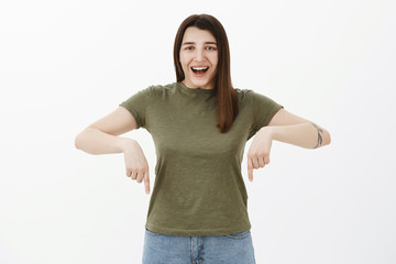 Happy and delighted bright optimistic female with brown hair and eyes in olive t-shirt smiling broadly and pointing up as pleasantly showing product, promoting copy space downwards over gray wall