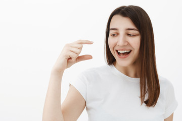 Close-up shot of amused and happy upbeat brunette in 20s laughing out loud and smiling delighted as shaping something tiny or small and looking and hand with grin over gray background