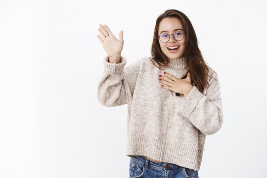 Girl Making Promise, Swearing Keep Secret Raising One Hand And Holding Arm On Heart As Making Pledge With Calm Happy Expression, Smiling And Gazing At Camera Kind And Pleased Over Gray Wall