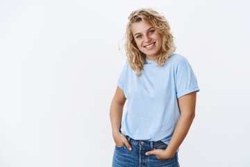 Pleasant and cute bright blonde female with blue eyes tilting head shy and grinning happily as...