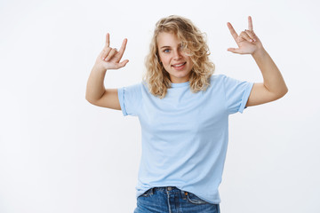 Fototapeta na wymiar Let roll. Cool and stylish carefree charismatic young 20s woman with blue eyes in t-shirt smiling having fun raising hands up with rock-n-roll gesture feeling awesome dancing and grinning at party