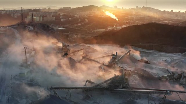 Flying sideways, aerial view of quarry with equipment and light smoke from it. Cityscape of Vladivostok is on the background. Evening, twilight
