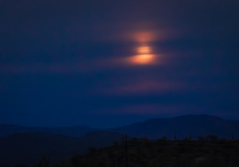 Full moon rising over a desert mountain in the Sonoran wilderness in southwest USA. The colorful full moon rises behind clouds in the night sky, giving the photograph texture in these photos 