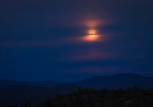 Full moon rising over a desert mountain in the Sonoran wilderness in southwest USA. The colorful full moon rises behind clouds in the night sky, giving the photograph texture in these photos 