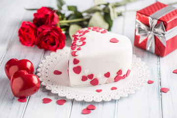 Obraz na płótnie Canvas Marzipan white cake in the shape of a heart with red hearts. As the decoration bouquet of red roses a gift from the ribbon. Wedding or valentines day concept.