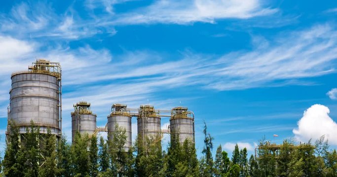 Passing cloud chemical powder silos behind row of tree with blue sky.
