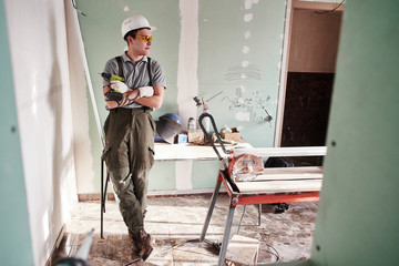 Room repair. Young builder in a helmet and overalls standing with folded hands on the background of the construction site