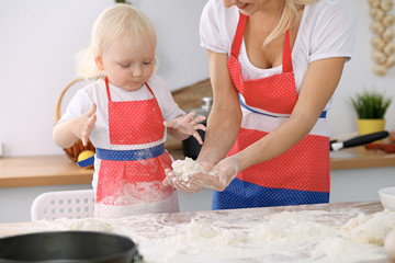 Little girl and her blonde mom in red aprons  playing and laughing while kneading the dough in kitchen. Homemade pastry for bread, pizza or bake cookies. Family fun and cooking concept